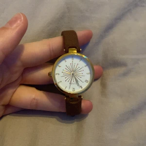 Guldig klocka med brun mockarem från Euro - Stilren klocka från Euro med rund vit urtavla, guldiga detaljer och ett unikt stjärnmönster i mitten. Klockan har en brun mockarem och spänne i guld. Perfekt accessoar för dig som gillar en klassisk men ändå cool look.