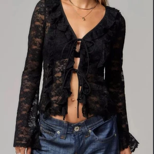Svart spetsblus med volang och snörning - Säljer en svart transparent spetsblus med långa ärmar, volangdetaljer längs v-ringningen och två snörningar framtill. Blusen är croppad och har en romantisk vibe, perfekt att styla med jeans eller kjol.