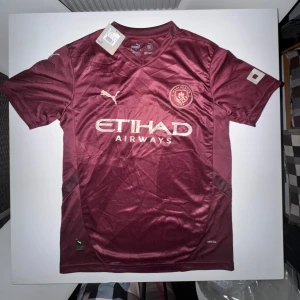 Puma x Manchester City fotbollströja  - Snygg vinröd Manchester City fotbollströja från Puma I STORLEK M! med Etihad Airways-tryck på bröstet. Tröjan har korta ärmar, V-ringning och är tillverkad i ett lätt, ventilerande material med DryCELL-teknologi. Perfekt för dig som vill sticka ut på planen eller läktaren.