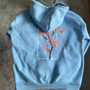 Blå hoodie med körsbärsblommor - Ljusblå hoodie med huva och snörning, fleecefodrad insida och stor körsbärsblomma-tryck på ryggen. Texten 'CHERRY TREE' och japanska tecken vid motivet. Perfekt för dig som gillar unika prints och mjuka material.