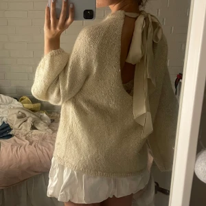 Beige stickad tröja med rosett i ryggen - Superfin beige stickad tröja med öppen rygg och stor rosett som knyts i nacken. Tröjan har vida ärmar och en mjuk, lite fluffig känsla. Perfekt för dig som gillar unika detaljer och en avslappnad men ändå söt stil.