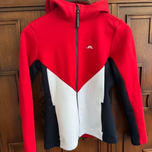Crystal Hood J.Lindeberg - Snygg hoodiejacka från J.Lindeberg i rött, vitt och svart. Jackan har hel dragkedja, huva och diskret logga på bröstet samt J.Lindeberg-tryck på huvan. Tillverkad i mjuk bomullsmix, perfekt för en sportig och avslappnad stil.
