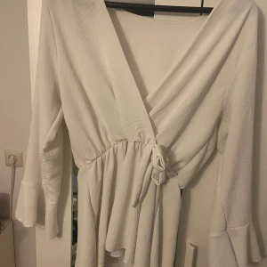 Vit omlottopp med volangärm - Supersnygg vit omlottopp med knyt i midjan och v-ringning. Toppen har långa ärmar med volangdetalj vid ärmslutet och ett flowigt fall. Perfekt för dig som gillar en feminin och trendig stil.
