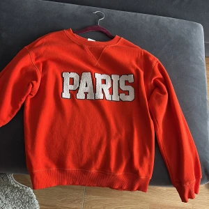 Orange sweatshirt med PARIS-tryck - Cool orange sweatshirt med stort vitt PARIS-tryck på bröstet. Klassisk rund halsringning och långa ärmar. Mjuk och skön bomullskänsla. 