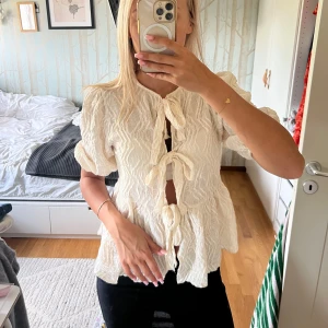 Vit blus med knyt från Gina Tricot - Supersöt vit blus från Gina Tricot med puffiga korta ärmar och tre knyt framtill. Blusen har en vågig, strukturerad yta och är lätt och luftig – perfekt för varmare dagar. Passformen är normal och materialet känns som bomull eller syntet.