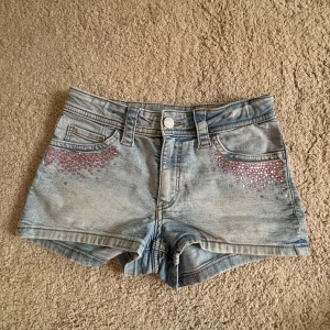 Jeansshorts med rosa stenar H&M - Snygga ljusblå jeansshorts från H&M med rosa glittrande stenar vid fickorna framtill. Klassisk femficksmodell med bälteshällor och knappstängning. Perfekta för sommaren och ger en cool vibe till din outfit.