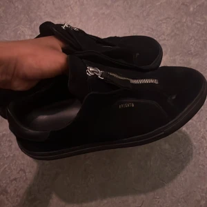 Svarta sneakers från Axel Arigato - Stilrena svarta sneakers från Axel Arigato i mocka med silverfärgad dragkedja på ovansidan. Skorna har låg profil, rund tå och diskret logga i guld på sidan. Perfekta för dig som gillar minimalistisk design och snygga detaljer.