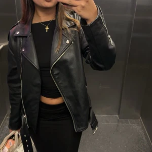 Svart skinnjacka med dragkedjor - Säljer en svart skinnjacka i klassisk bikerstil med silvriga dragkedjor och bälte i midjan. Jackan har breda slag, långa ärmar och coola detaljer som ger en edgy vibe. Perfekt att slänga över en hoodie eller t-shirt för en snygg streetlook.