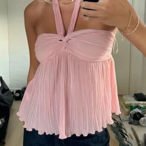 Rosa halterneck topp från Zara - Supersöt ljusrosa halterneck topp med plisserat tyg och twistad detalj framtill från Zara. Toppen har en flowy passform och breda axelband som knyts i nacken. Perfekt för varma dagar när du vill ha något luftigt och trendigt. Lappen sitter kvar.