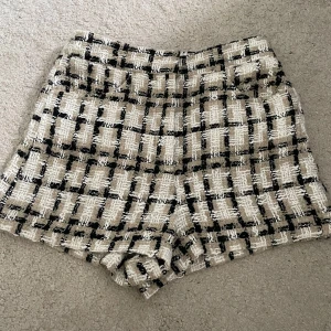 Rutiga tweedshorts från Zara - Snygga shorts i tweed med rutigt mönster i beige, svart och vitt från Zara. De har en hög midja och en lite boxig passform som ger en cool och trendig vibe. Perfekta att styla med en enkel topp eller kavaj för en chic look.