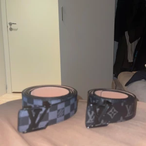 Louis Vuitton svarta bälten med mönster - Två snygga Louis Vuitton-bälten i svart med ikoniska LV-spännen. Ena bältet har klassiskt rutigt mönster i svart och grått, det andra har LV-monogram och blommor i grått på svart botten. Båda är i skinn och har en modern vibe som lyfter vilken outfit som helst. En för 250
