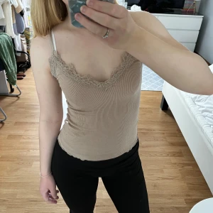 Beige ribbad linnetopp med spets - Supersöt beige linnetopp med ribbad struktur och fina spetsdetaljer längs v-ringningen. Toppens smala axelband och figurnära passform gör den perfekt för en chill och stilren look. Toppen är från New Yorker och har används ett fåtal gånger, men är fortfarande i fint skick 🥰