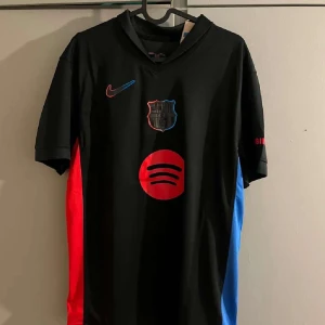 FC Barcelona svart Nike fotbollströja - Snygg svart FC Barcelona fotbollströja från Nike med klubbmärke och Spotify-logga på bröstet. Röda och blå detaljer längs sidorna och röd Ambilight tv-tryck på ärmen. Tröjan är kortärmad och tillverkad i ett lätt, andningsbart material perfekt för träning och sommaren.