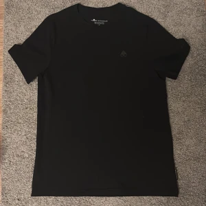 Svart t-shirt från Moose Knuckles - En stilren svart t-shirt från Moose Knuckles i 100% bomull. Klassisk passform med korta ärmar och diskret logga på bröstet samt tryckt varumärke på ärmen. Perfekt för dig som gillar minimalistisk och clean stil. Äkta T-shirt aldrig använd pga att den e stor