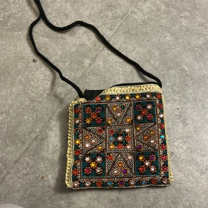 Broderad axelväska med pärlor och dragkedja - Unik axelväska med broderade mönster och färgglada pärlor i orange, blått, vitt och guld. Väskan har svart tyg, guldiga detaljer och stängs med dragkedja. Perfekt för dig som gillar boho-stil och vill sticka ut med accessoarer.