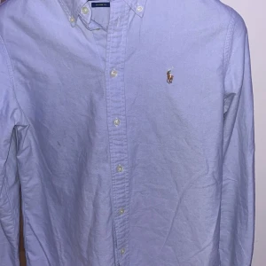 Ljusblå skjorta från Ralph Lauren - Klassisk ljusblå skjorta från Ralph Lauren med broderad logga på bröstet. Skjortan har knappar hela vägen, button-down krage och lång ärm. Perfekt för en clean och stilren look. 