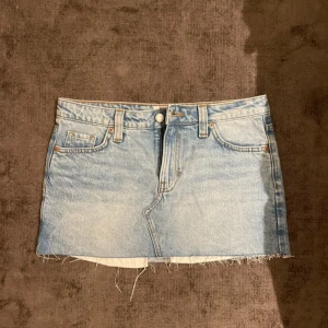 Ljusblå jeanskjol från Monki - Säljer en ljusblå jeanskjol från Monki i klassisk femficksmodell. Kjolen är kort med rå, fransig fåll och har bälteshällor samt knapp- och dragkedjestängning. Aldrig använd