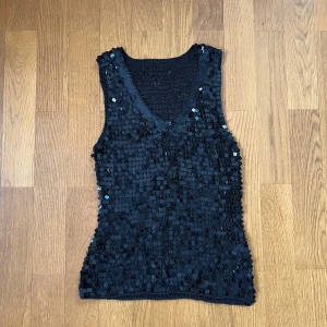 Svart paljett-topp med v-ringning - Säljer en svart ärmlös topp täckt med glittrande paljetter. Toppen har v-ringning framtill och är figurnära i modellen. Perfekt för dig som vill sticka ut med en riktigt glittrig look. Materialet är syntet och paljetterna ger en cool och festlig känsla.