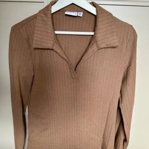 Ribbad brun/beige långärmad tröja från VILA  - Säljer en ribbad brun långärmad tröja från Vila med krage och v-ringad öppning. Tröjan har en slim passform och är tillverkad i mjukt material som sitter snyggt på kroppen. Perfekt för dig som gillar stilrena och trendiga basplagg.