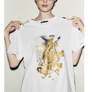 Vit t-shirt med guldtryck från Zara - Vit t-shirt från Zara med coolt tryck framtill i svartvitt och stora detaljer i glittrigt guld. Klassisk passform med korta ärmar och rund hals. Slutsåld på hemsidan. Aldrig använd. 