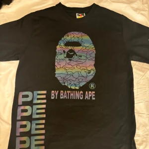 Svart BAPE t-shirt med regnbågstryck - Svart t-shirt från BAPE med ikoniskt ape-head tryck i regnbågsfärger på bröstet och texten 'BY BATHING APE'. Flerfärgat tryck längs sidan och bak, samt en unik etikett på ärmen. Klassisk passform och rund hals. Perfekt statement-plagg för streetwear. Storlek large men känns mer som en medium