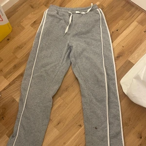 Grå mjukisbyxor med vita ränder - Snygga grå mjukisbyxor med vita ränder längs sidorna och snörning i midjan. Perfekta för chill dagar eller när du vill vara bekväm men ändå se fräsch ut. Klassisk passform och mjukt material.