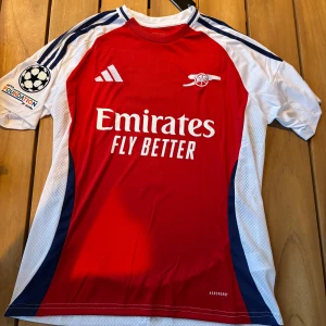 Arsenal Adidas matchtröja 24/25 - Arsenal fotbollströja med Saka 7 på ryggen. Röd front med vita och marinblå detaljer, Adidas-logga och klubbmärke broderat. Kortärmad modell i lätt funktionsmaterial (AEROREADY). Klassisk design med vita axelpartier och blå ränder på ärmarna.