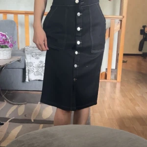 Svart jeanskjol med knappar - Svart jeanskjol med hög midja och stora framfickor. Kjolen har vita kontrastsömmar och knappar hela vägen framtill. Snygg och enkel att styla med crop top eller stickad tröja.