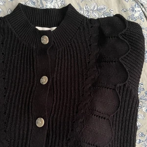 Svart stickad kofta med knappar - Säljer en jätte gullig svart stickad kofta med snygga detaljer längs axlarna och framstycket. Koftan har stora, ljusa knappar och ett ribbat mönster som ger en cool struktur. Perfekt för lager-på-lager och passar till allt i garderoben.
