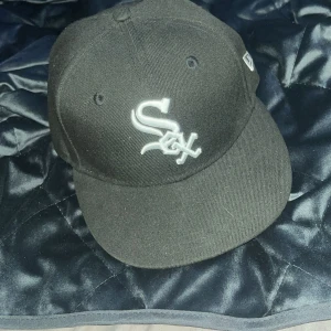 Svart New Era 59FIFTY White Sox keps - Snygg svart 59FIFTY keps från New Era med Chicago White Sox-logga broderad i vitt framtill. Klassisk basebollmodell med böjd skärm och MLB-logga bak. Perfekt för dig som gillar streetwear och sportig stil.