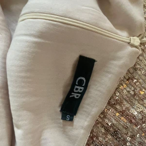 Glittrig champagnefärgad klänning CBR - Supersnygg långklänning från CBR i champagnefärg med massor av paljetter. Klänningen har offshoulder-modell och trekvartsärm, perfekt för dig som vill sticka ut. Den är figurnära och har dragkedja i sidan för enkel påklädning.