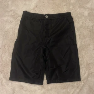 Svarta shorts från Manfinity - Stilrena svarta shorts från Manfinity med klassisk passform och midja. De har bälteshällor, knapp och dragkedja framtill samt sidfickor. Materialet känns lätt och svalt, perfekt för varma dagar. Enkla att matcha med det mesta och passar dig som gillar en clean look.