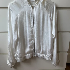 Vi, tunn bomberjacka från Kappahl - Snygg vit bomberjacka från Kappahl med dragkedja framtill och elastiska muddar vid ärmslut och midja. Jackan har en clean look och är tillverkad i ett lätt, glansigt material som ger en fräsch vibe. Jackan är mycket tunn och luftig. 