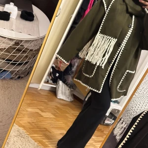 Olivgrön kappa med fransar och kontrastsöm - Säljer en olivgrön kappa med vita kontrastsömmar och långa fransar framtill. Jackan har stora fickor, knappar och en oversized passform. Perfekt statement-plagg för dig som vill sticka ut med coola detaljer och unik look.