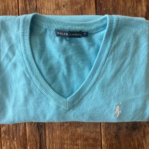 Turkos v-ringad tröja från Ralph Lauren - Snygg turkos v-ringad tröja från Ralph Lauren med klassisk broderad logga på bröstet. Tröjan har långa ärmar och ribbade muddar vid ärmslut och nederkant. Perfekt för dig som gillar stilrena och fräscha plagg med premiumkänsla.