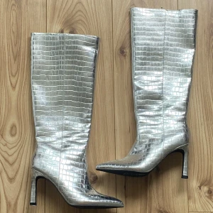 Silvriga höga boots med krokoprägling - Statement boots i metallic-silver med krokodilpräglat mönster och spetsig tå. Höga skaft och smal klack ger en riktigt edgy vibe. Perfekta för dig som vill sticka ut och addera lite futuristisk känsla till outfiten.