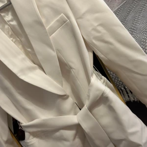 Vit kavaj med bälte från Zara - Snygg vit kavaj från Zara med breda slag och knytbälte i midjan. Kavajen har en stilren design med fickdetaljer och är helfodrad för en lyxig känsla. Perfekt för dig som vill ha en fräsch och modern look.
