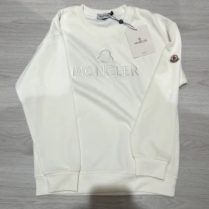 Vit Moncler sweatshirt med broderad logga - Snygg vit sweatshirt från Moncler med broderad logga framtill och klassisk rund hals. Tröjan har ribbade muddar vid ärmslut och nederkant samt Moncler-märke på ärmen. Perfekt för dig som gillar stilrena och exklusiva plagg.