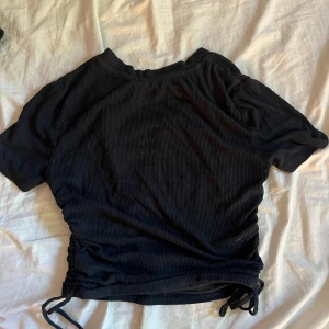 Svart ribbad topp med snörning - Svart ribbad topp med korta ärmar och snygg snörningsdetalj i sidan. Den är jätte fin och sitter snyggt