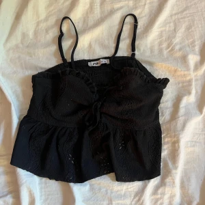 Svart linnetopp med spets och knyt - Supersöt svart linnetopp med smala axelband, volangkant upptill och dekorativ knytning framtill. Toppen har fina spetsdetaljer. Jätte gullig