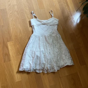 Vit spetsklänning från Gina Tricot XS - Supersöt vit kort klänning från Gina Tricot i storlek XS. Klänningen har tunna axelband, spets med blommigt mönster och en liten rosett framtill. Perfekt för sommarens alla tillfällen när du vill känna dig extra fin.