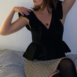 Svart volangblus med v-ringning - Supersnygg svart blus med djup v-ringning och volangdetaljer på axlarna. Modellen har en markerad midja och lätt utsvängd nederdel som ger en feminin siluett. Perfekt att styla med smycken och kjol för en trendig look.