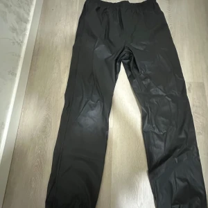 Svarta täckbyxor med resårmidja regn byxor  - Svarta täckbyxor med enkel design och resår i midjan. Perfekta för kalla dagar eller utomhusaktiviteter. Materialet är syntetiskt och byxorna har en rak passform som ger rörelsefrihet.