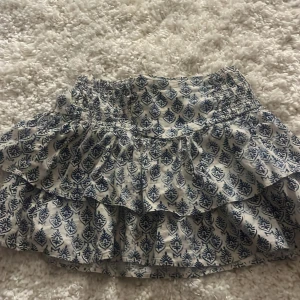 Vit och blå volangkjol från Zara - Supersöt kort volangkjol från Zara i vitt med blått mönster. Kjolen har resår i midjan och dubbla volanger som ger en flowig känsla. Perfekt för varma dagar och lätt att matcha med en enkel topp. Den har en liten brun fläck som knappt syns på baksidan av kjolen, fläcken går inte bort men den syns knapp när den är på! På sista bilden ser man fläcken.💕