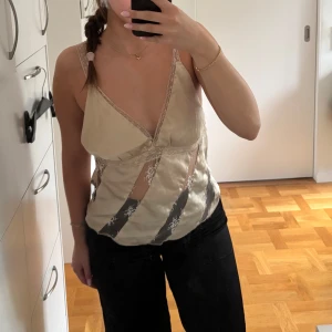 Beige spetslinnetopp - Säljer en beige linnetopp med svarta diagonala spetsdetaljer och tunna spetsband. Toppen har v-ringning och är ärmlös med öppen rygg. Materialet är glansigt och känns lyxigt. Aldrig använd! 