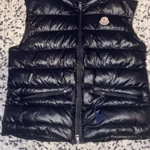 Svart dunväst från Moncler - Snygg svart dunväst från Moncler med glansig finish och quiltad design. Västen har två dragkedjefickor framtill, hög krage och klassisk Moncler-logga på bröstet. Perfekt att slänga över en hoodie för en cool look.