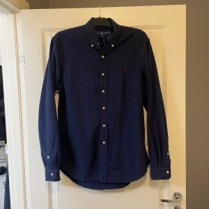Mörkblå skjorta från Ralph Lauren - Klassisk mörkblå skjorta från Ralph Lauren med button-down krage och vita knappar. Skjortan har lång ärm och den ikoniska röda loggan broderad på bröstet. Tillverkad i mjuk bomull, perfekt för en clean och stilren look.