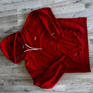 Röd hoodie från FILA med snörning - Snygg röd hoodie från FILA med vit snörning i huvan och diskret logga på bröstet och ärmen. Klassisk passform med långa ärmar och känguruficka framtill. Perfekt för dig som gillar sportig och avslappnad stil.