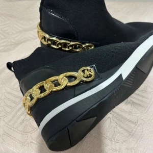 Svarta Michael Kors boots med guldkedja - Svarta boots från Michael Kors i stickad textil med chunky vit och svart sula. Snygg guldfärgad kedjedetalj med MK-logga runt hälen ger en lyxig vibe. Skorna har rund tå och platt sula, perfekt för dig som gillar statement-detaljer.