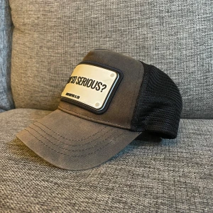 Svart truckerkeps John Hatter & Co - Cool svart truckerkeps från John Hatter & Co med vit patch framtill där det står 'WHY SO SERIOUS?'. Kepsen har böjd skärm och mesh baktill för extra ventilation. Perfekt för dig som vill sticka ut med en statement-accessoar.